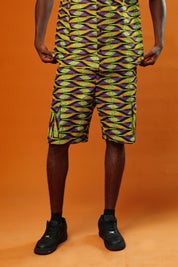Man wearing Aztec green men’s shorts made from African print cotton fabric with a yellow, black, and purple geometric pattern. The shorts are knee-length with a relaxed fit and side cargo pockets. Paired with a matching shirt, black trainers, and sunglasses. Orange background.