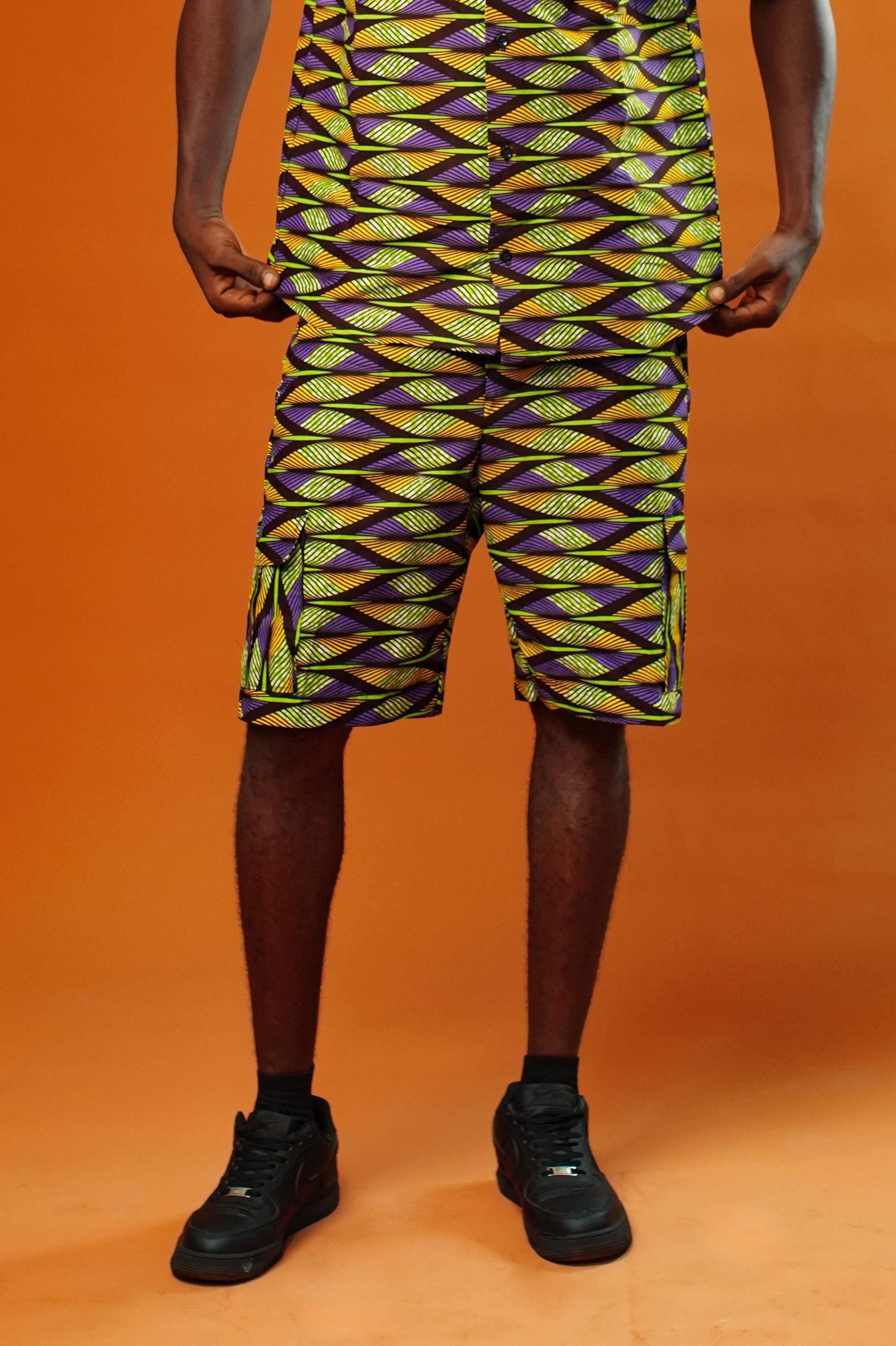 Man wearing Aztec green men’s shorts made from African print cotton fabric with a yellow, black, and purple geometric pattern. The shorts are knee-length with a relaxed fit and side cargo pockets. Paired with a matching shirt, black trainers, and sunglasses. Orange background.