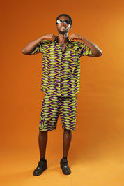 A man strikes a confident pose against a bold orange background, wearing a matching two-piece outfit made from vibrant African wax print fabric. The short-sleeved button-up shirt and shorts feature a striking geometric pattern in neon green, purple, and black.