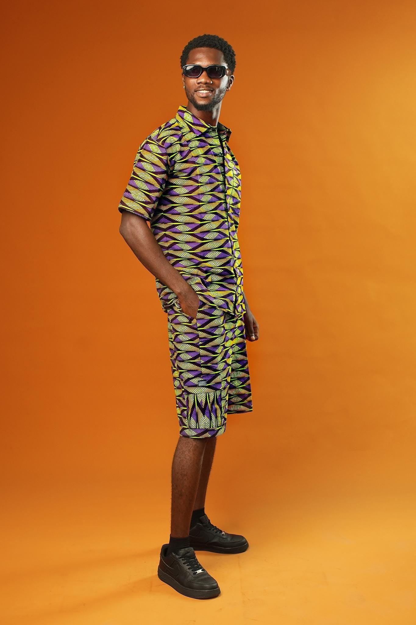 A man strikes a confident pose against a bold orange background, wearing a matching two-piece outfit made from vibrant African wax print fabric. The short-sleeved button-up shirt and shorts feature a striking geometric pattern in neon green, purple, and black.