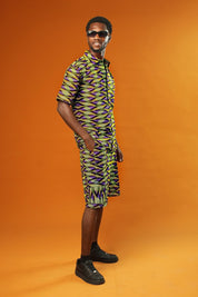 A man strikes a confident pose against a bold orange background, wearing a matching two-piece outfit made from vibrant African wax print fabric. The short-sleeved button-up shirt and shorts feature a striking geometric pattern in neon green, purple, and black.