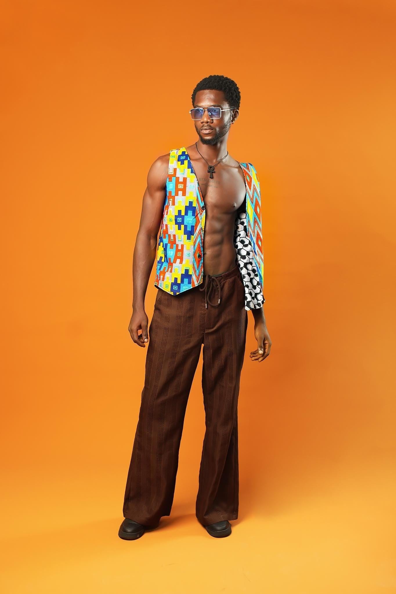 A man stands in front of an orange background wearing a Kente Reversible Men Waistcoat with bright Aztec African print in red, yellow, blue, and green. The vest is sleeveless, open at the chest, and made from Ankara cotton. He is also wearing brown drawstring trousers, a black ankh necklace, and blue-tinted sunglasses.