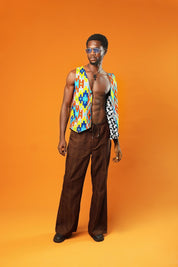 A man stands in front of an orange background wearing a Kente Reversible Men Waistcoat with bright Aztec African print in red, yellow, blue, and green. The vest is sleeveless, open at the chest, and made from Ankara cotton. He is also wearing brown drawstring trousers, a black ankh necklace, and blue-tinted sunglasses.