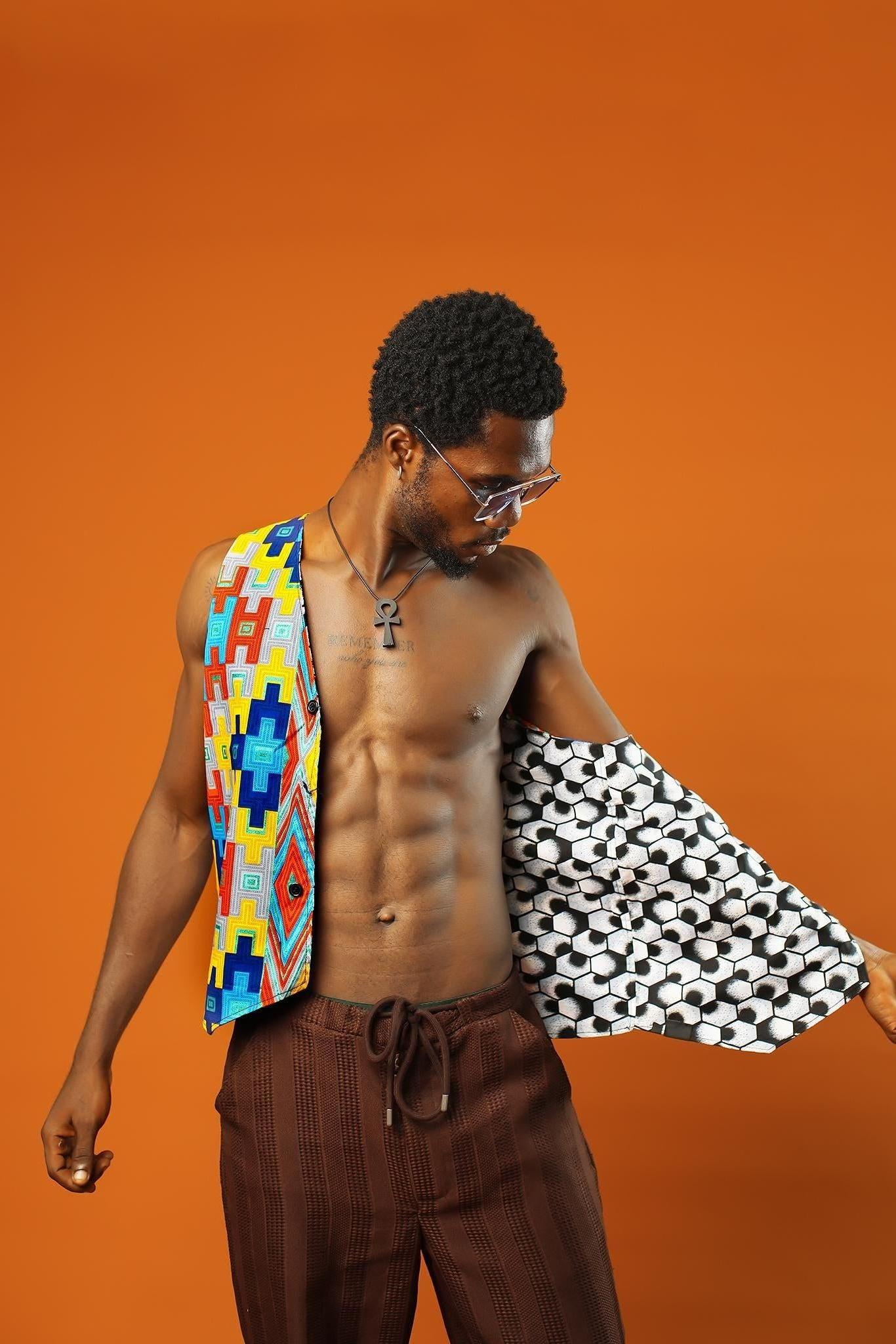 A man stands in front of an orange background wearing a Kente Reversible Men Waistcoat with bright Aztec African print in red, yellow, blue, and green. The vest is sleeveless, open at the chest, and made from Ankara cotton. He is also wearing brown drawstring trousers, a black ankh necklace, and blue-tinted sunglasses.