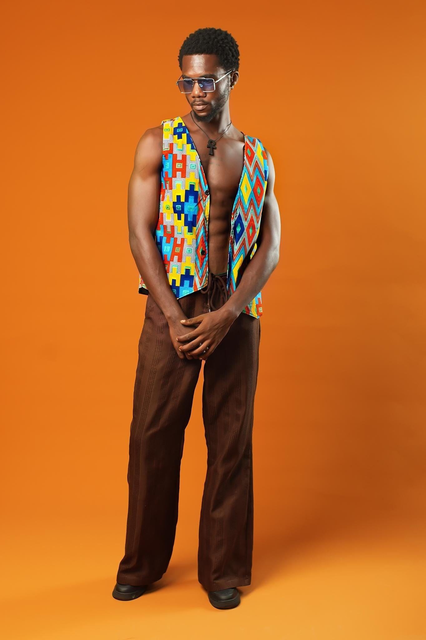 A man stands in front of an orange background wearing a Kente Reversible Men Waistcoat with bright Aztec African print in red, yellow, blue, and green. The vest is sleeveless, open at the chest, and made from Ankara cotton. He is also wearing brown drawstring trousers, a black ankh necklace, and blue-tinted sunglasses.