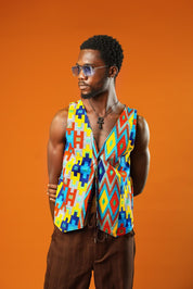 A man poses stylishly against an orange background wearing a bold Kente Reversible Men Waistcoat made from vibrant Ankara cotton. The Aztec African print vest features bright geometric patterns in yellow, red, blue, and orange. A standout festival outfit for men, perfect for hippie clothing lovers or fans of yellow kawaii and Afro-inspired fashion.