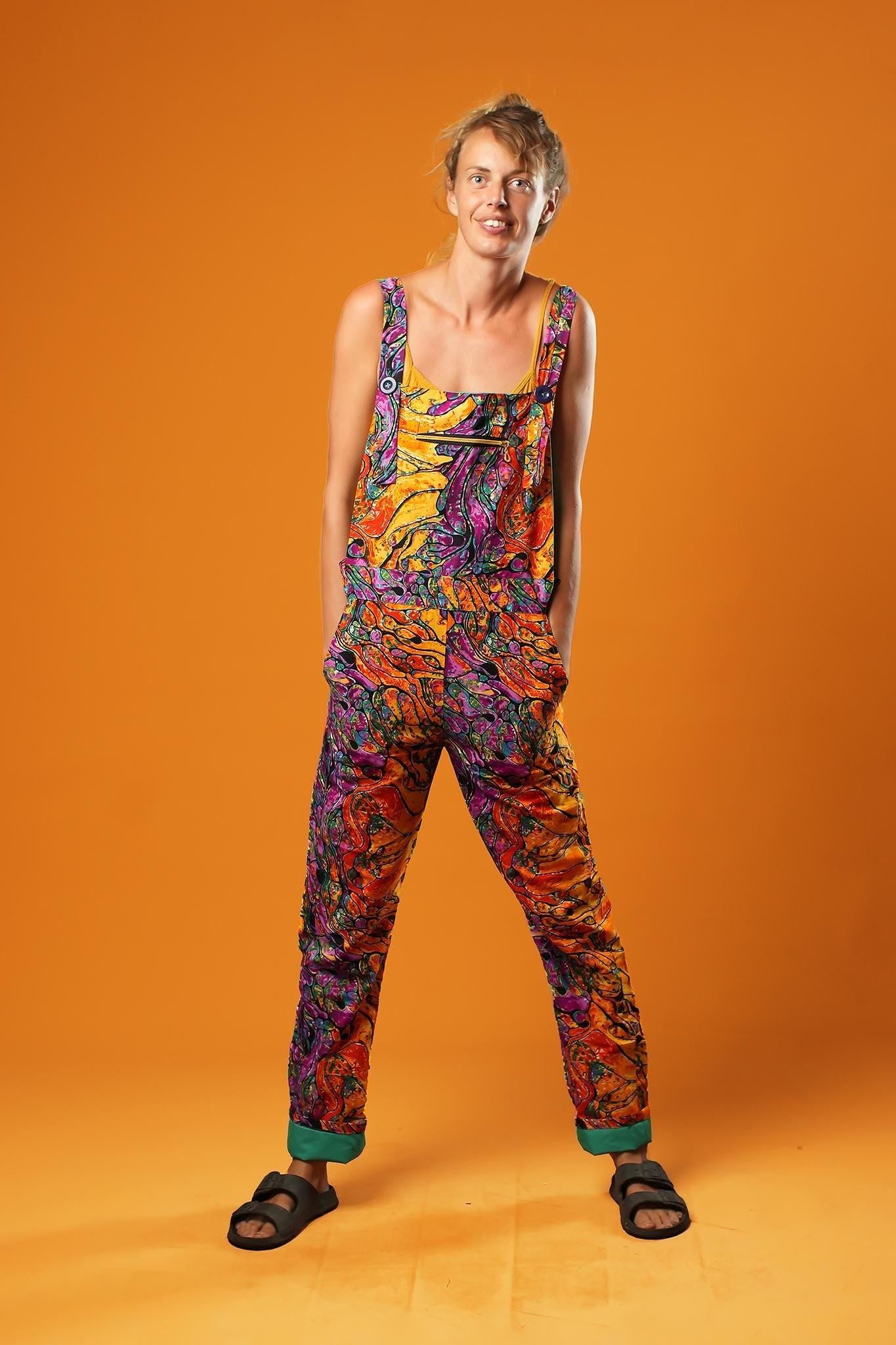 Woman standing against an orange background wearing colourful unisex dungarees. The dungarees feature a vibrant orange, purple, green, and yellow abstract African print with teal ankle cuffs and adjustable straps