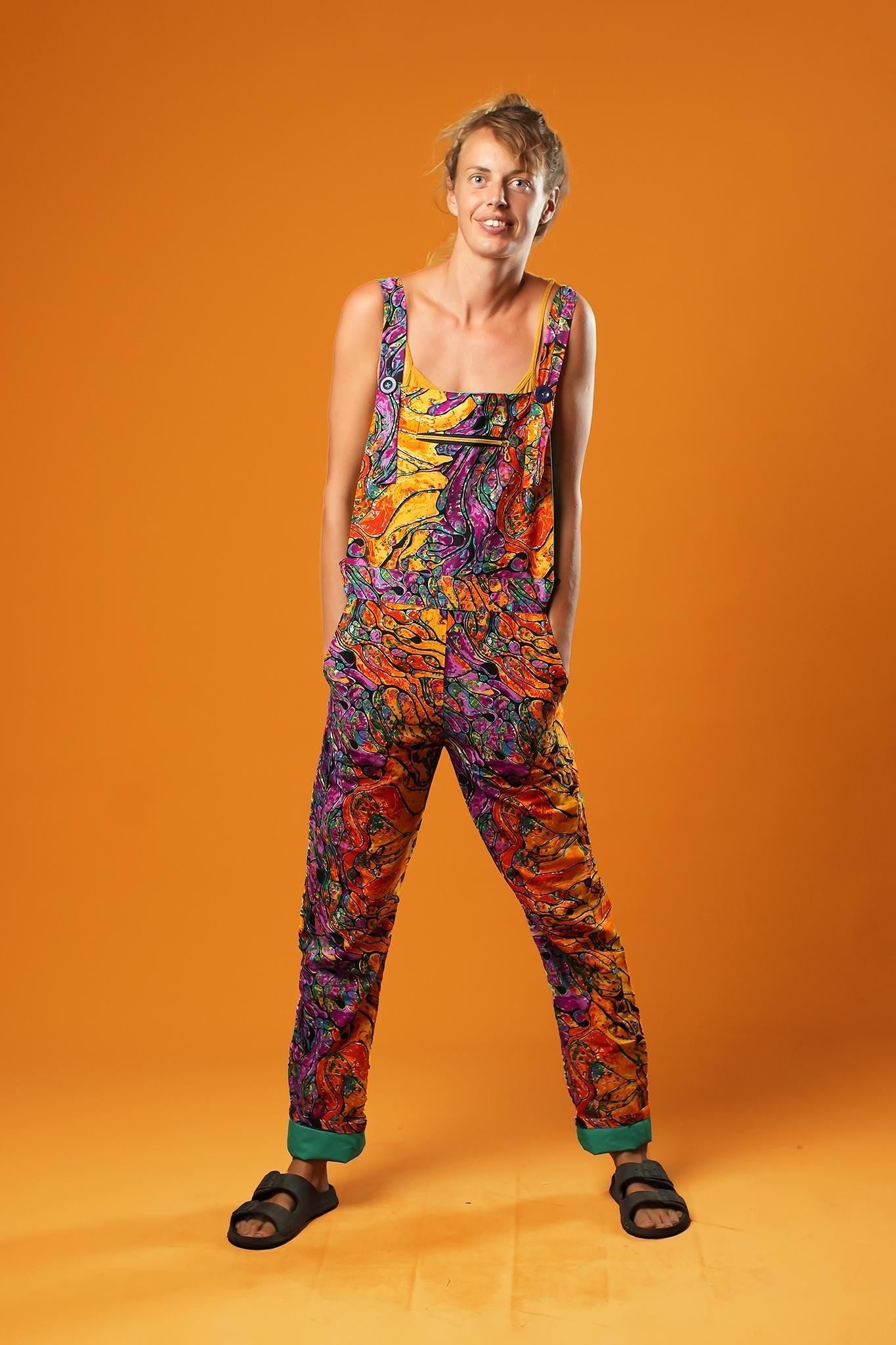 Woman standing against an orange background wearing colourful unisex dungarees. The dungarees feature a vibrant orange, purple, green, and yellow abstract African print with teal ankle cuffs and adjustable straps