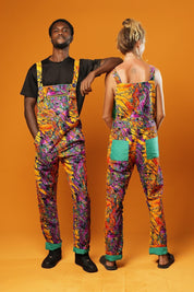 Man and woman standing against an orange background wearing matching colourful unisex dungarees. The dungarees feature a vibrant orange, purple, green, and yellow abstract African print with green ankle cuffs and adjustable straps