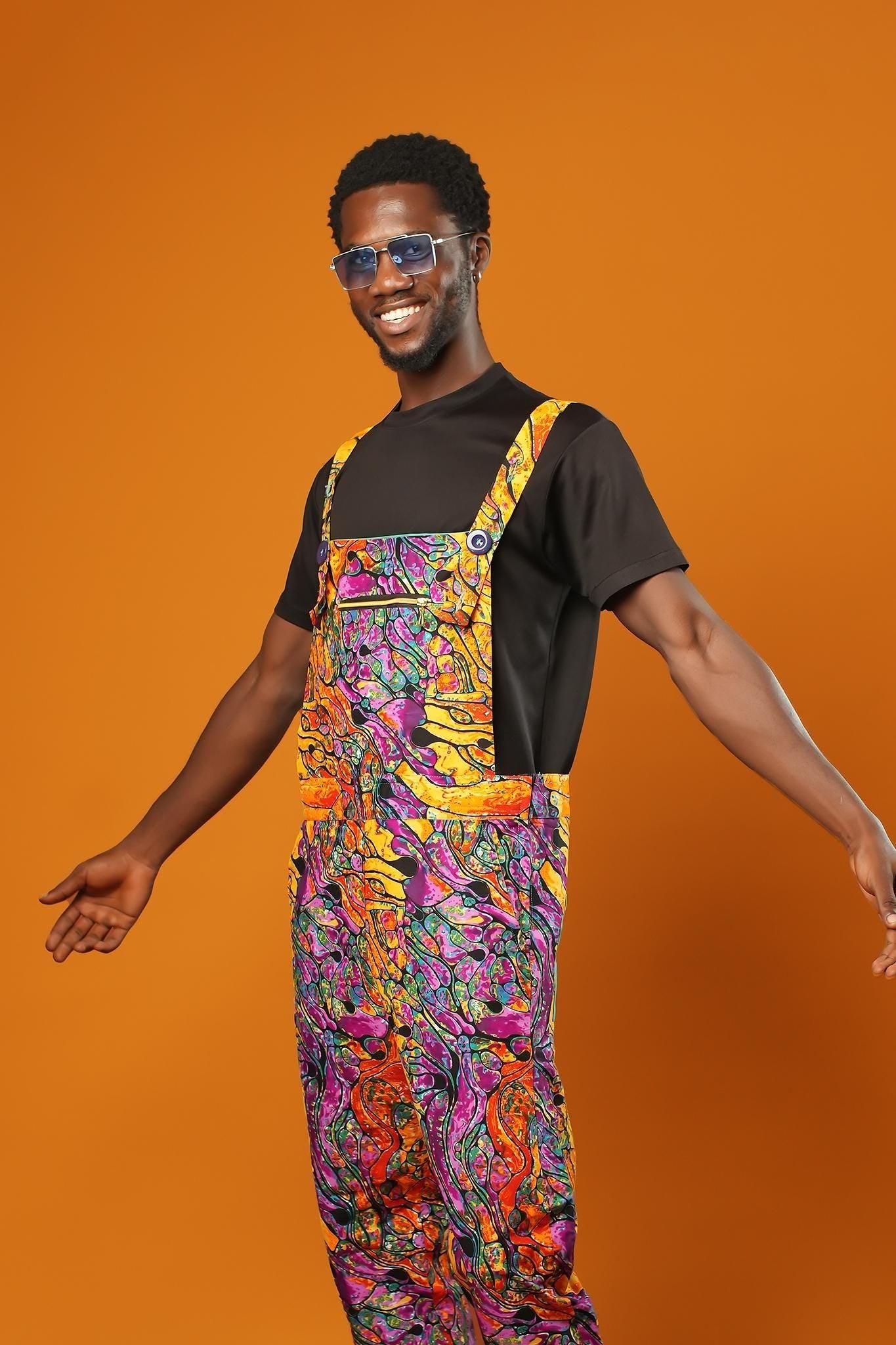 Man standing against an orange background wearing colourful unisex dungarees. The dungarees feature a vibrant orange, purple, green, and yellow abstract African print with teal ankle cuffs and adjustable straps