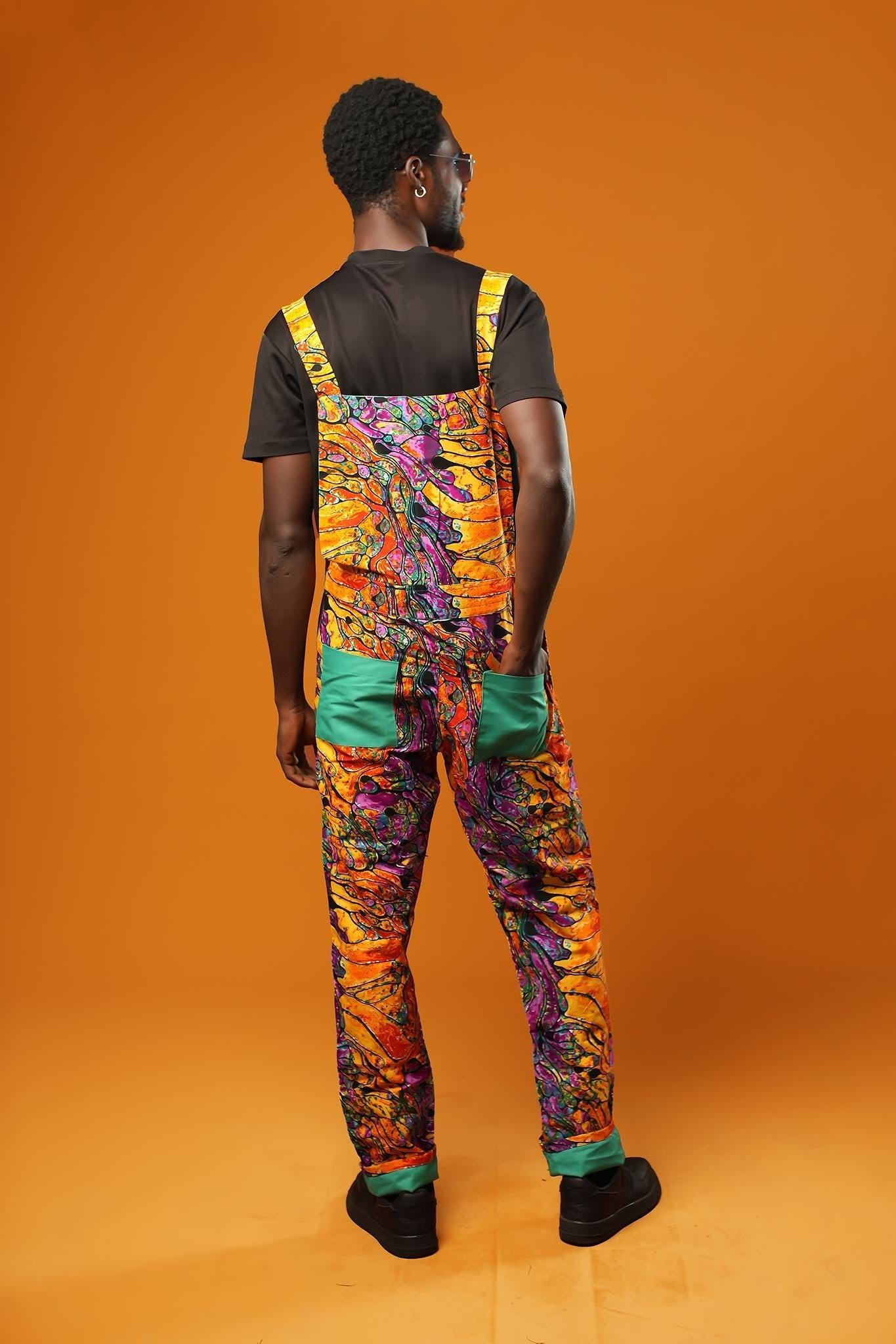 Woman standing against an orange background wearing colourful unisex dungarees. The dungarees feature a vibrant orange, purple, green, and yellow abstract African print with teal ankle cuffs and adjustable straps