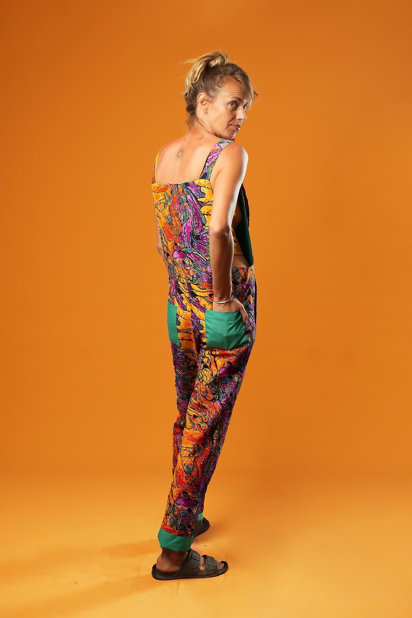 Woman standing against an orange background wearing colourful unisex dungarees. The dungarees feature a vibrant orange, purple, green, and yellow abstract African print with teal ankle cuffs and adjustable straps