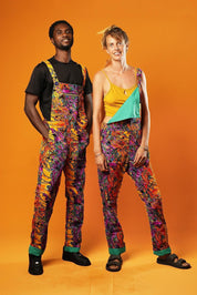 Man and woman standing against an orange background wearing matching colourful unisex dungarees. The dungarees feature a vibrant orange, purple, green, and yellow abstract African print with green ankle cuffs and adjustable straps