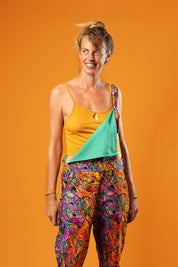 Woman standing against an orange background wearing colourful unisex dungarees. The dungarees feature a vibrant orange, purple, green, and yellow abstract African print.