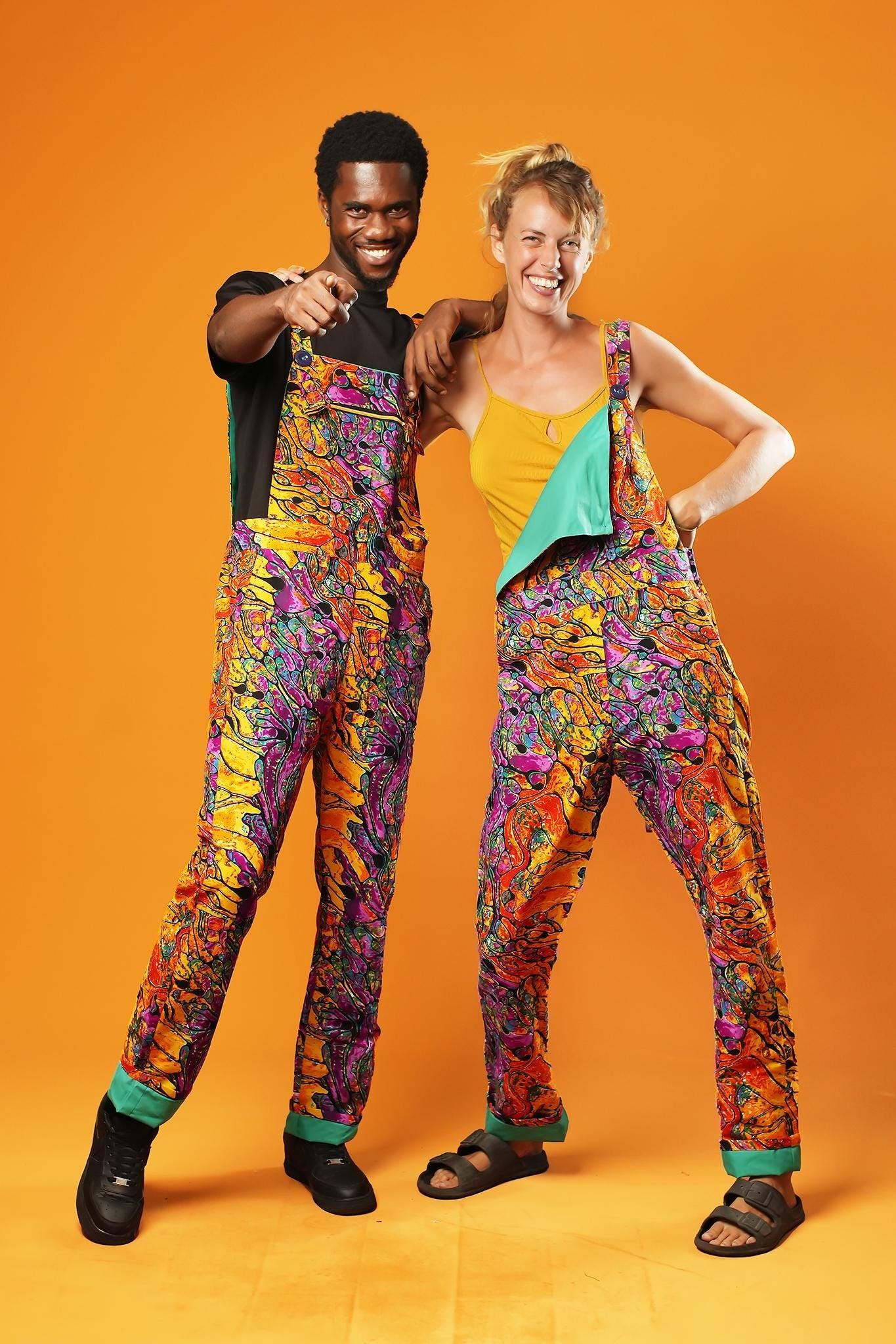 Man and woman standing against an orange background wearing matching colourful unisex dungarees. The dungarees feature a vibrant orange, purple, green, and yellow abstract African print with green ankle cuffs and adjustable straps