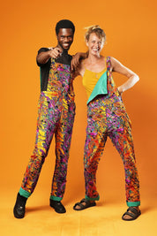 Man and woman standing against an orange background wearing matching colourful unisex dungarees. The dungarees feature a vibrant orange, purple, green, and yellow abstract African print with green ankle cuffs and adjustable straps