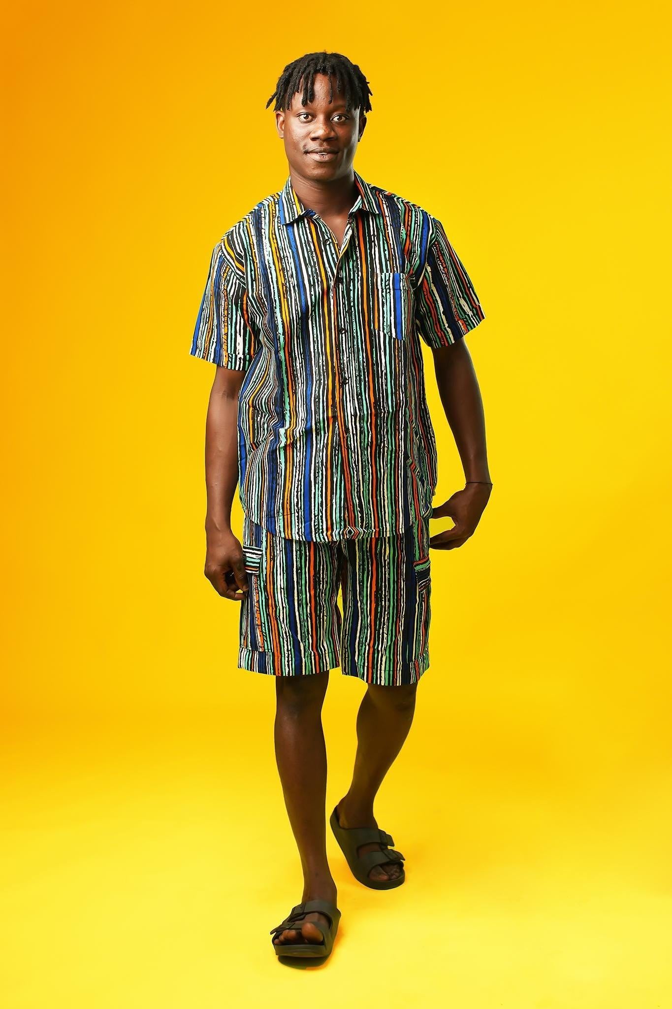Man wearing African print shorts with large patch cargo pockets, made from blue striped African print cotton fabric with orange, green, black, and white lines. The shorts are paired with a matching short-sleeve shirt. Yellow background.