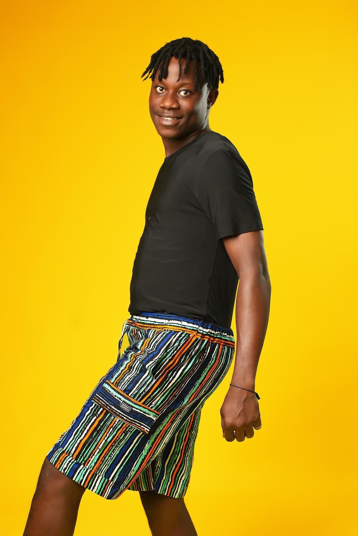 Man wearing African print shorts with large patch cargo pockets, made from blue striped African print cotton fabric with orange, green, black, and white lines. The shorts are paired with a matching short-sleeve shirt. Yellow background.