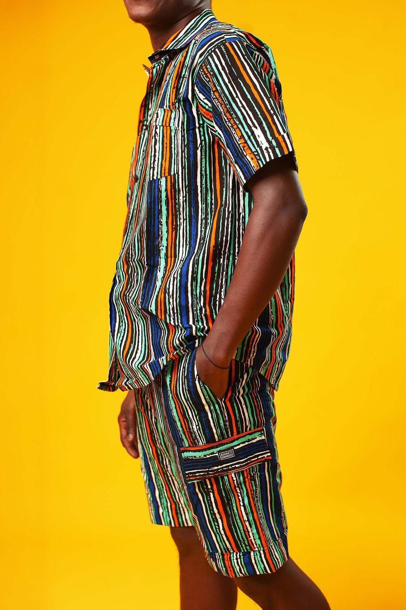 Close-up side view of a man wearing striped African print cargo shorts in green, blue, orange, black, and white. The shorts have a relaxed fit with large side pockets and a visible Ashanti Empress label. He has one hand in the pocket and is wearing a matching short-sleeve shirt. The background is bright yellow. This is part of a summer festival outfit and funky men’s clothing set.