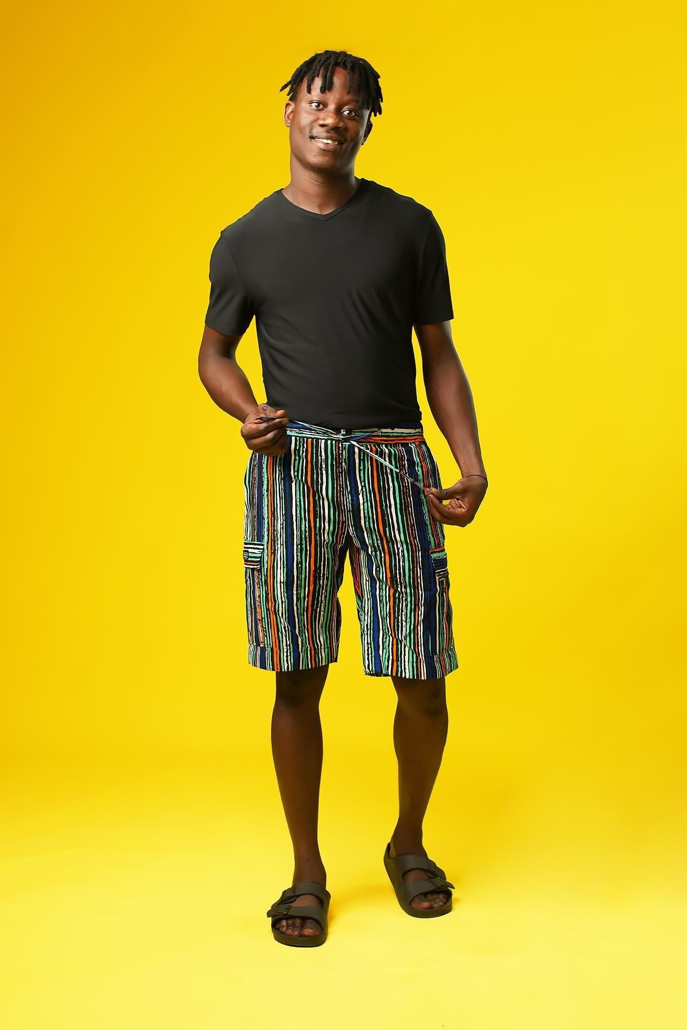 Man wearing African print shorts with large patch cargo pockets, made from blue striped African print cotton fabric with orange, green, black, and white lines. The shorts are paired with a matching short-sleeve shirt. Yellow background.