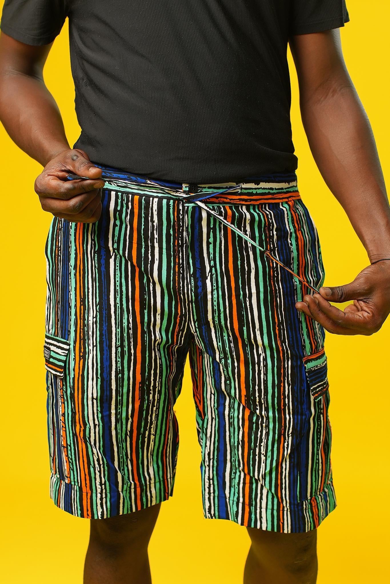 Man wearing African print shorts with large patch cargo pockets, made from blue striped African print cotton fabric with orange, green, black, and white lines. The shorts are paired with a matching short-sleeve shirt. Yellow background.