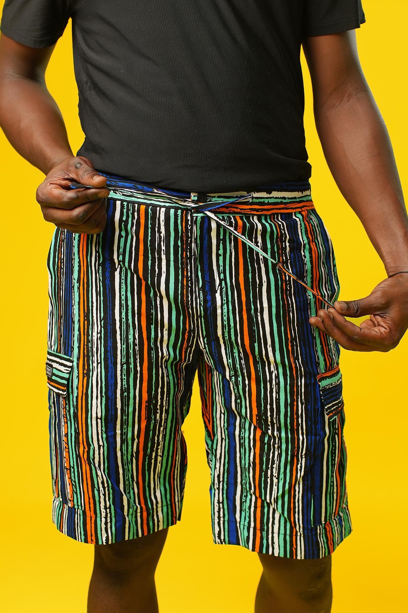 Man wearing African print shorts with large patch cargo pockets, made from blue striped African print cotton fabric with orange, green, black, and white lines. The shorts are paired with a matching short-sleeve shirt. Yellow background.