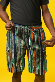 Man wearing African print shorts with large patch cargo pockets, made from blue striped African print cotton fabric with orange, green, black, and white lines. The shorts are paired with a matching short-sleeve shirt. Yellow background.
