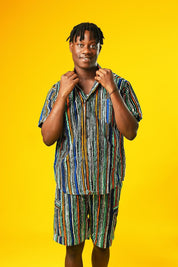 A man walks confidently against a vibrant yellow background wearing a stylish matching two-piece outfit. The set features a short-sleeved button-up shirt and shorts made from a multicoloured striped African print fabric with vertical lines in blue, green, orange, black, and white. He pairs the look with black slide sandals, creating a relaxed yet bold summer-ready vibe.
