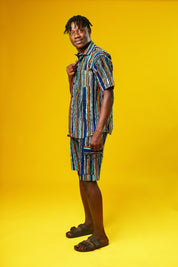 A man walks confidently against a vibrant yellow background wearing a stylish matching two-piece outfit. The set features a short-sleeved button-up shirt and shorts made from a multicoloured striped African print fabric with vertical lines in blue, green, orange, black, and white. He pairs the look with black slide sandals, creating a relaxed yet bold summer-ready vibe.