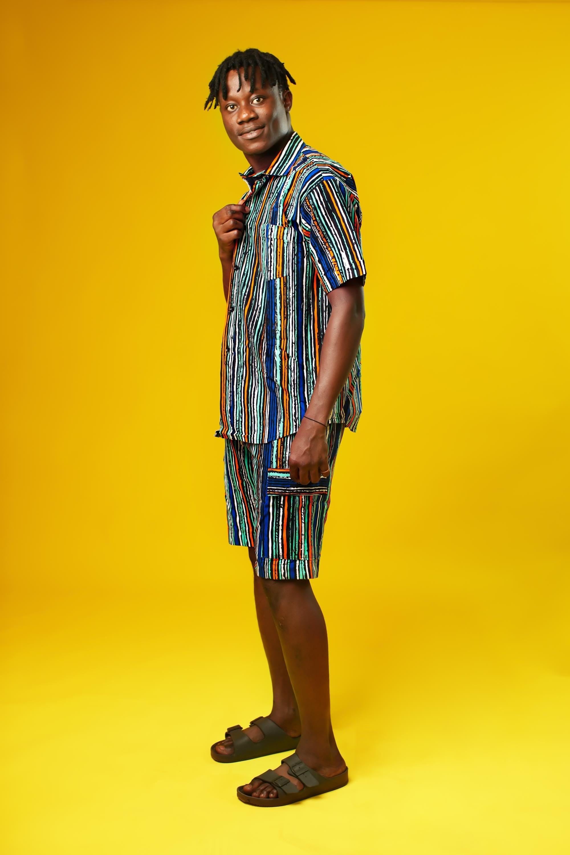 A man walks confidently against a vibrant yellow background wearing a stylish matching two-piece outfit. The set features a short-sleeved button-up shirt and shorts made from a multicoloured striped African print fabric with vertical lines in blue, green, orange, black, and white. He pairs the look with black slide sandals, creating a relaxed yet bold summer-ready vibe.