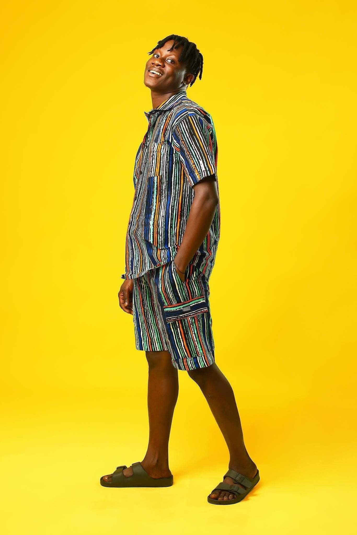 A man walks confidently against a vibrant yellow background wearing a stylish matching two-piece outfit. The set features a short-sleeved button-up shirt and shorts made from a multicoloured striped African print fabric with vertical lines in blue, green, orange, black, and white. He pairs the look with black slide sandals, creating a relaxed yet bold summer-ready vibe.