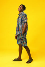 A man walks confidently against a vibrant yellow background wearing a stylish matching two-piece outfit. The set features a short-sleeved button-up shirt and shorts made from a multicoloured striped African print fabric with vertical lines in blue, green, orange, black, and white. He pairs the look with black slide sandals, creating a relaxed yet bold summer-ready vibe.