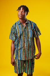 A man walks confidently against a vibrant yellow background wearing a stylish matching two-piece outfit. The set features a short-sleeved button-up shirt and shorts made from a multicoloured striped African print fabric with vertical lines in blue, green, orange, black, and white. He pairs the look with black slide sandals, creating a relaxed yet bold summer-ready vibe.