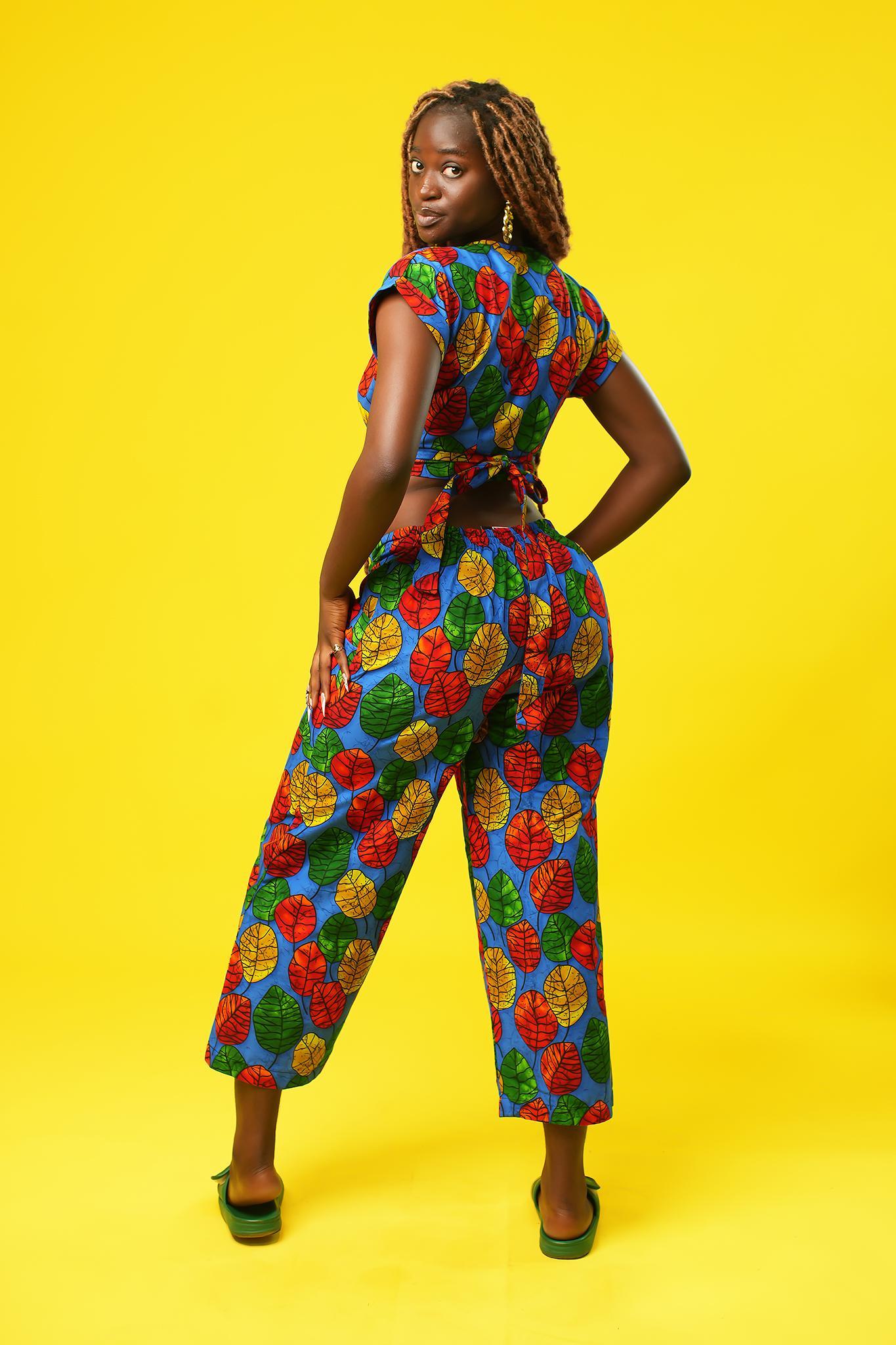 Woman wearing wide leg palazzo pants made from colourful African wax print cotton with a red, green, yellow, and blue leaf pattern. The cropped trousers have a high waist, relaxed fit, and side pockets, styled as part of a matching festival outfit.