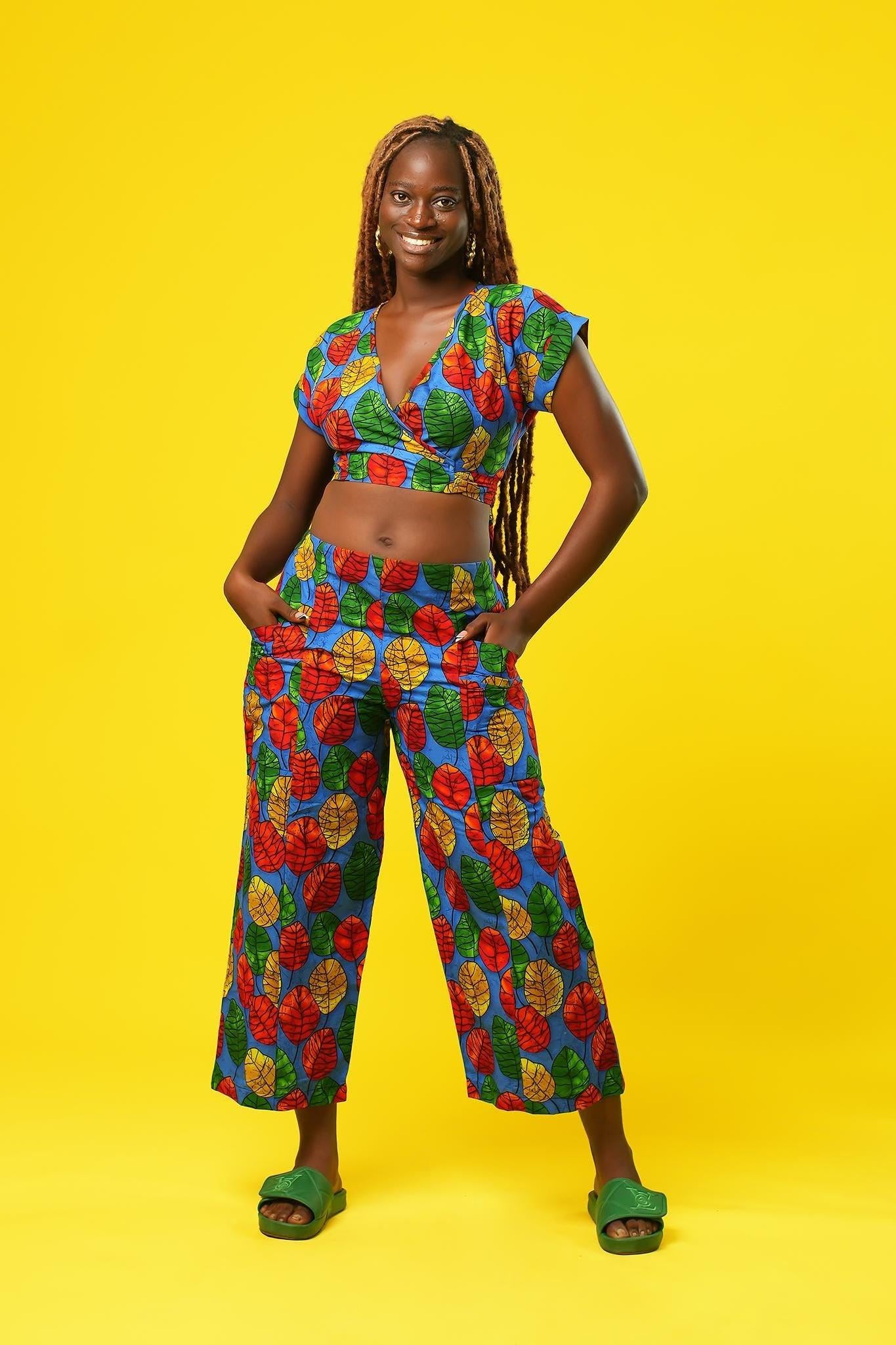 Woman wearing a colourful African print wrap top with a V-neck and short sleeves, made from bright blue fabric featuring red, yellow, and green leaf patterns. The top has a cropped fit and ties at the waist.