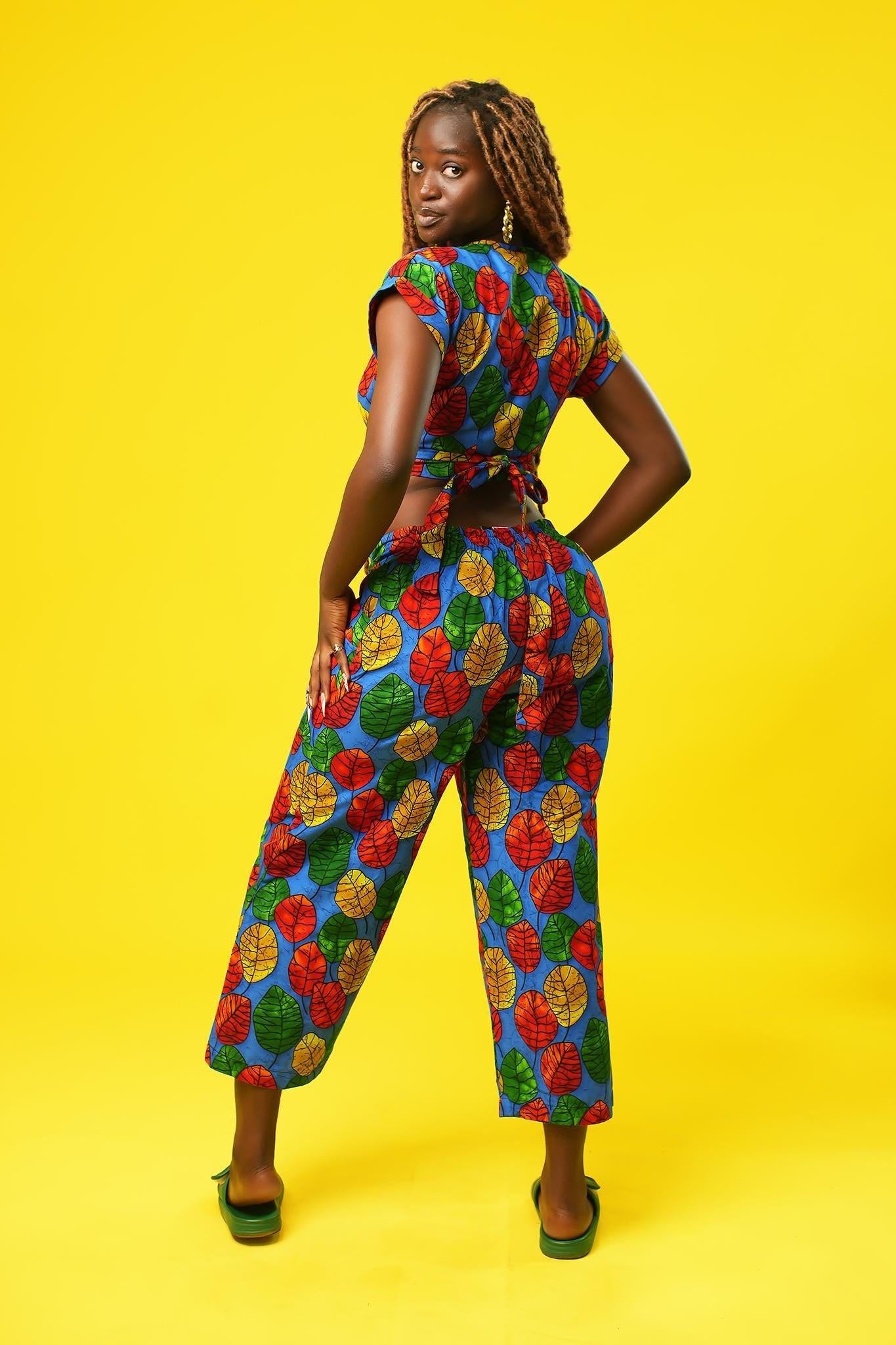 Woman wearing a colourful African print wrap top with a V-neck and short sleeves, made from bright blue fabric featuring red, yellow, and green leaf patterns. The top has a cropped fit and ties at the waist.