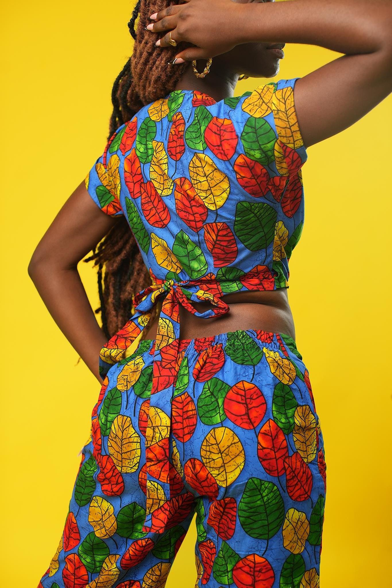 Woman wearing a colourful African print wrap top with a V-neck and short sleeves, made from bright blue fabric featuring red, yellow, and green leaf patterns. The top has a cropped fit and ties at the waist.