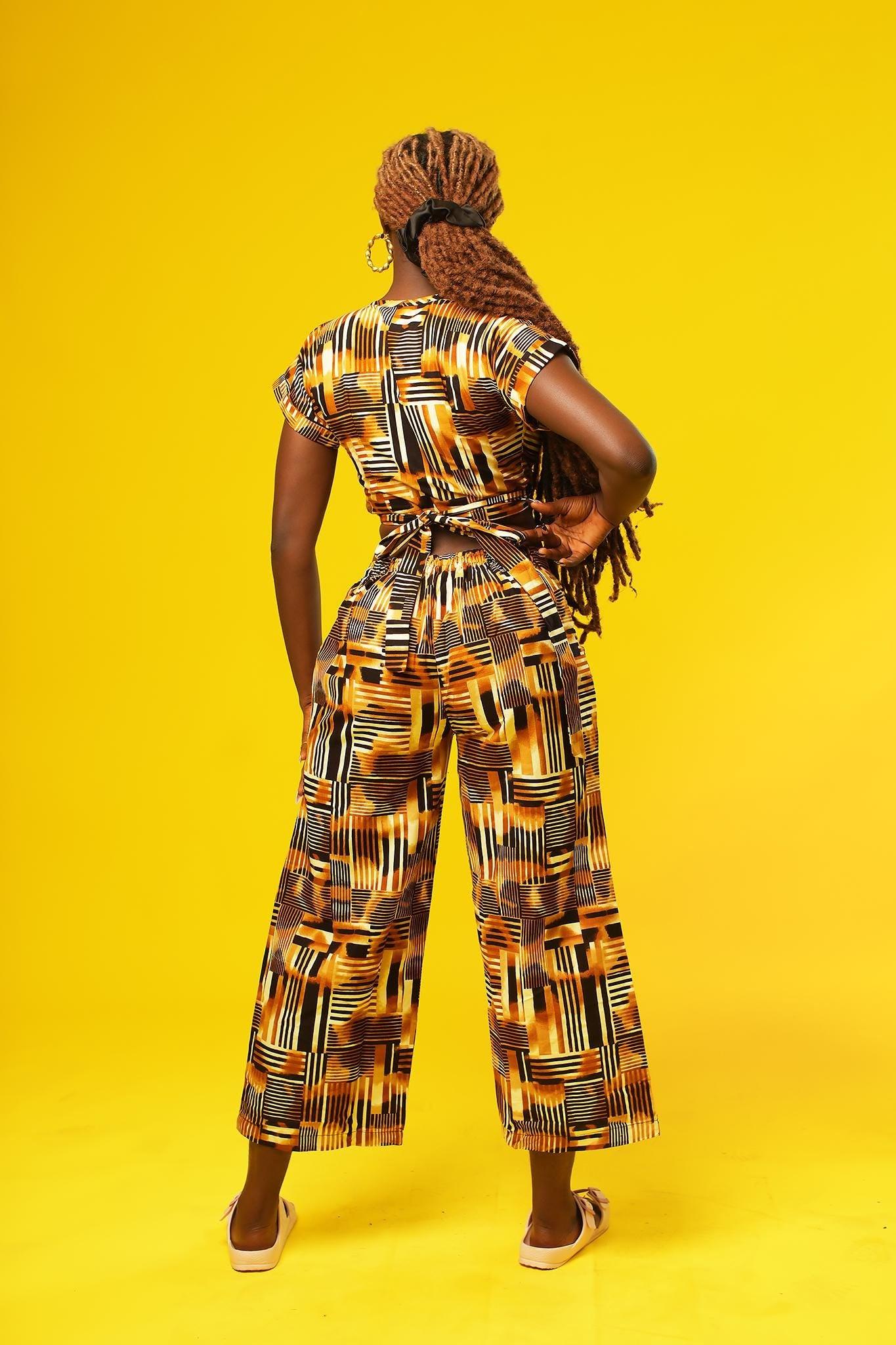 Woman wearing wide leg African palazzo pants with a geometric brown, yellow, orange, and black print. The cropped trousers are made from lightweight cotton, styled as part of a festival outfit with a matching wrap crop top.