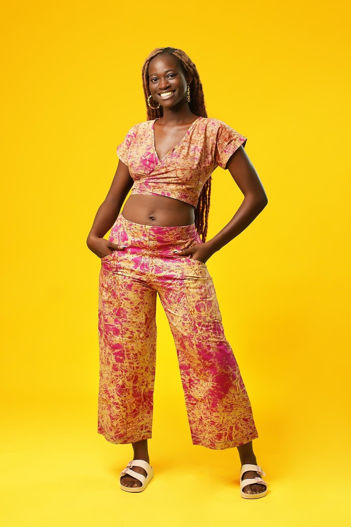 Woman wearing a pink and cream batik wrap crop top with short sleeves and a V-neck, made from African cotton fabric. The top features a fitted, wrap-around design and is paired with matching wide-leg trousers.