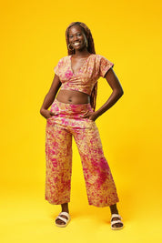 Woman wearing a pink and cream batik wrap crop top with short sleeves and a V-neck, made from African cotton fabric. The top features a fitted, wrap-around design and is paired with matching wide-leg trousers.