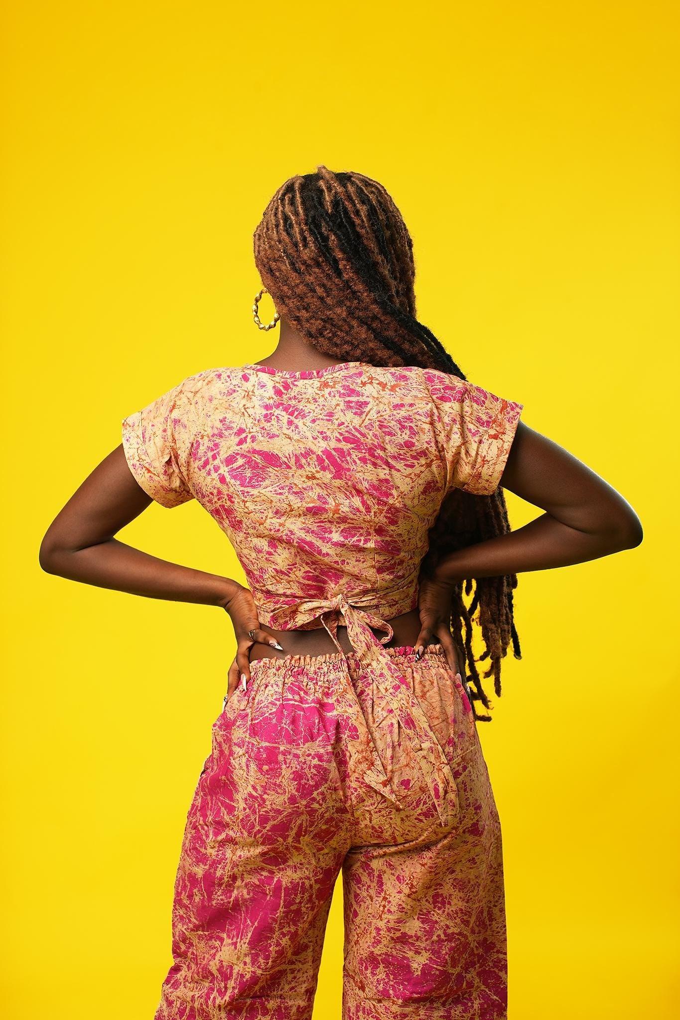 Woman wearing a pink and cream batik wrap crop top with short sleeves and a V-neck, made from African cotton fabric. The top features a fitted, wrap-around design and is paired with matching wide-leg trousers.