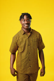 A man poses against a vibrant yellow background wearing a coordinated two-piece outfit made from a rich mustard yellow and black African print fabric. The set includes a short-sleeved button-up shirt and relaxed drawstring trousers featuring a bold geometric pattern.