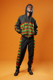 Man standing against an orange background wearing black tracksuit trousers with colourful Kente African print panels down the sides. The trousers have a relaxed fit with elastic cuffs at the ankles and a drawstring waist.