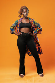 Woman wearing a colourful tie-dye African print midi kimono against an orange background. The unisex kimono features a vibrant pattern with shades of orange, purple, turquoise, and yellow, open at the front, with side pockets and loose sleeves.