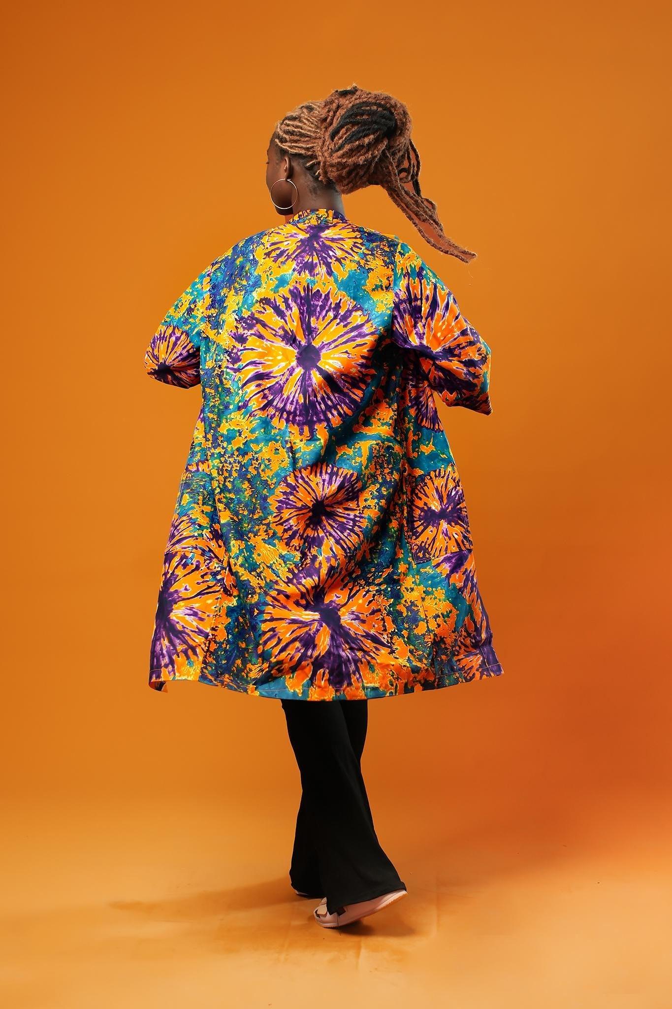 Woman wearing a colourful tie-dye African print midi kimono against an orange background. The unisex kimono features a vibrant pattern with shades of orange, purple, turquoise, and yellow, open at the front, with side pockets and loose sleeves.