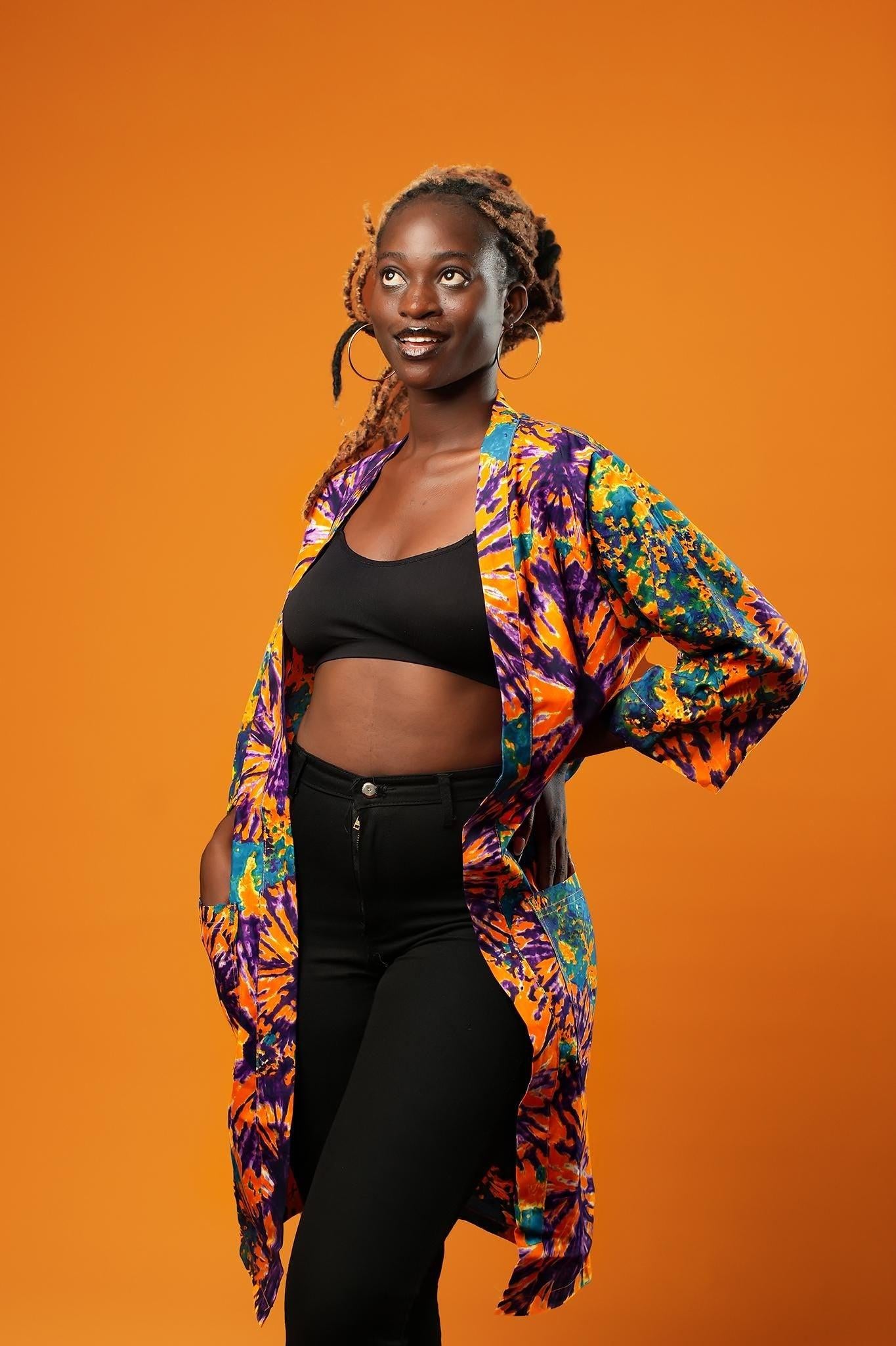 Woman wearing a colourful tie-dye African print midi kimono against an orange background. The unisex kimono features a vibrant pattern with shades of orange, purple, turquoise, and yellow, open at the front, with side pockets and loose sleeves.