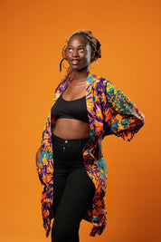 Woman wearing a colourful tie-dye African print midi kimono against an orange background. The unisex kimono features a vibrant pattern with shades of orange, purple, turquoise, and yellow, open at the front, with side pockets and loose sleeves.