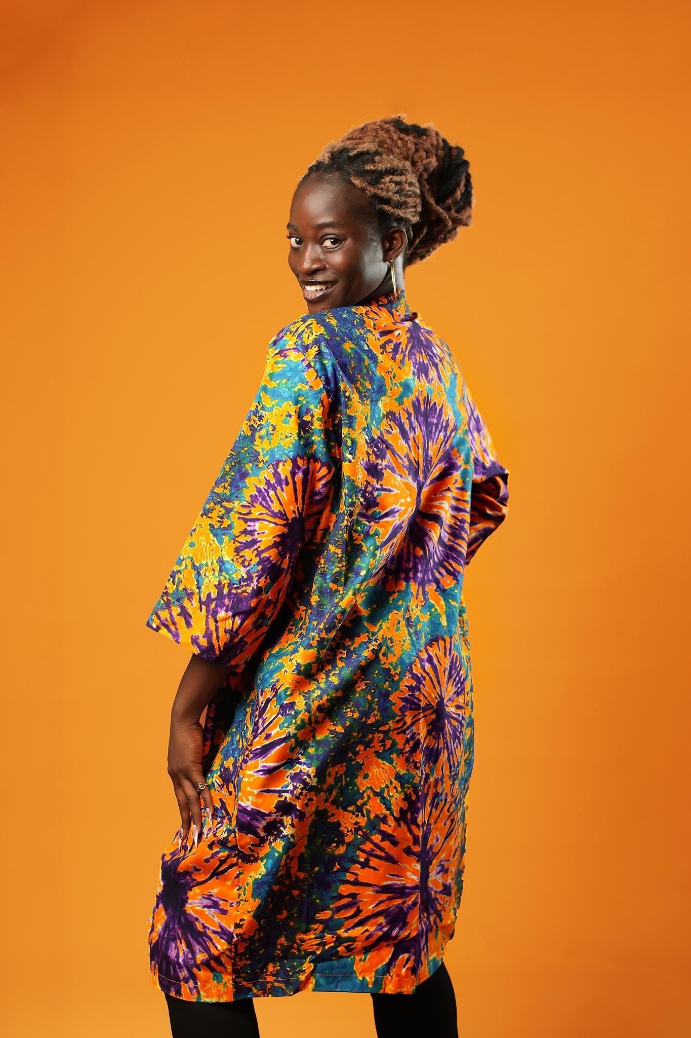 Woman wearing a colourful tie-dye African print midi kimono against an orange background. The unisex kimono features a vibrant pattern with shades of orange, purple, turquoise, and yellow, open at the front, with side pockets and loose sleeves.