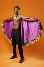 Man standing against an orange background wearing a purple festival cape with a colourful Kente African print trim. The cape ties at the neck with matching Kente fabric.