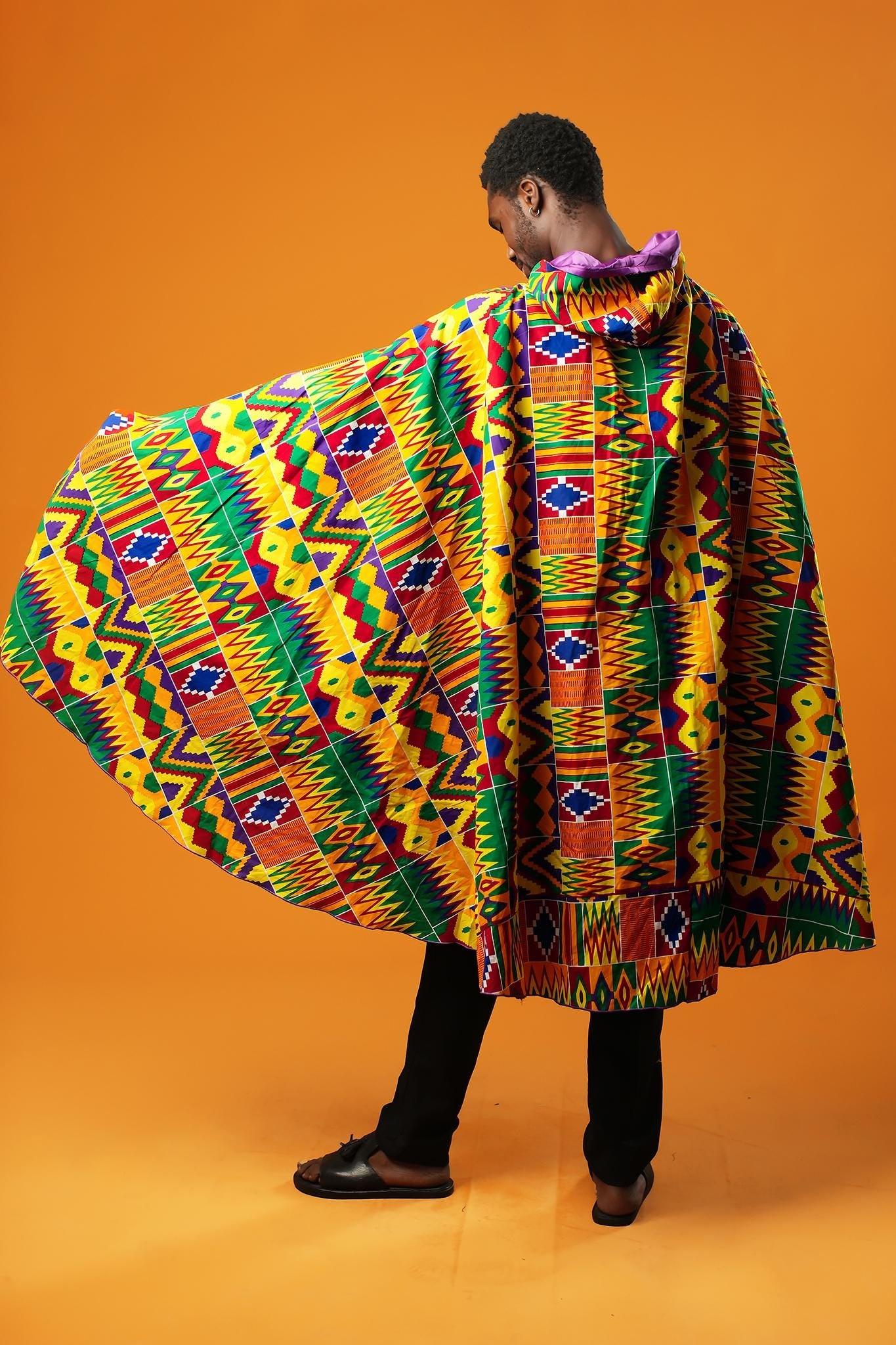 Man standing against an orange background wearing a purple festival cape with a colourful Kente African print trim. The cape ties at the neck with matching Kente fabric.